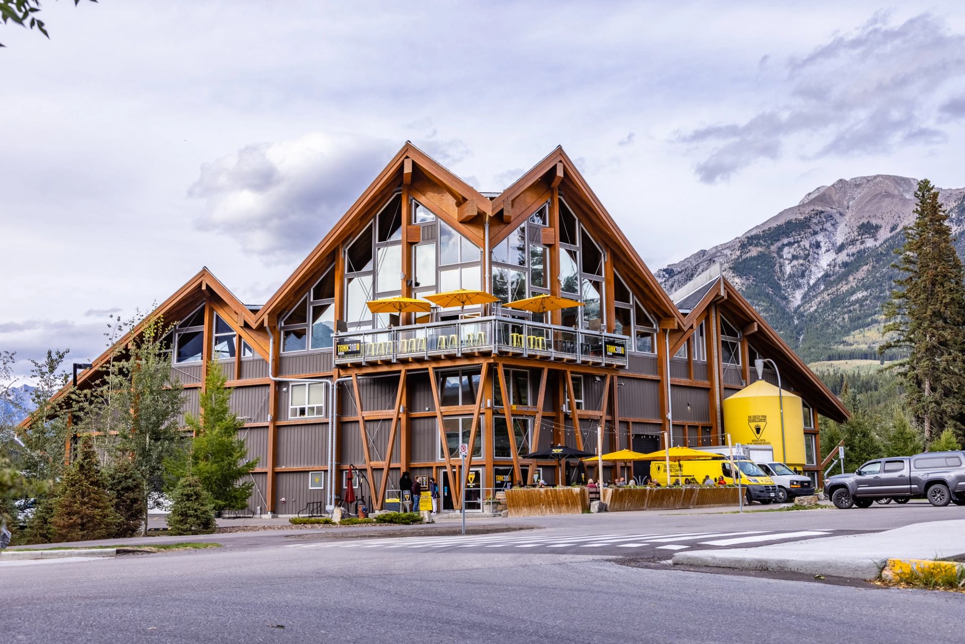 Wood and glass lodge-style building with yellow patio umbrellas, vehicles and mountains behind.