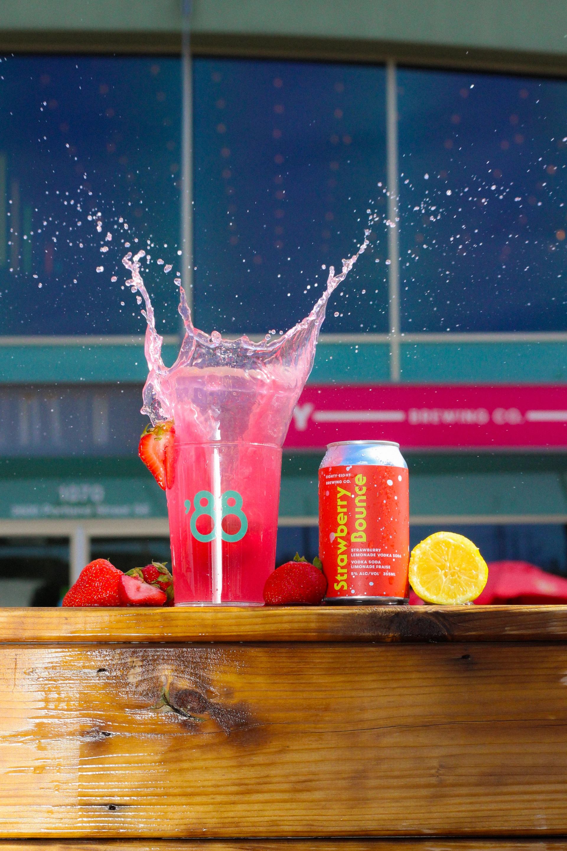 Splashing pink drink with fruit and a can of Eighty-Eight Brewing beer on a wooden table.