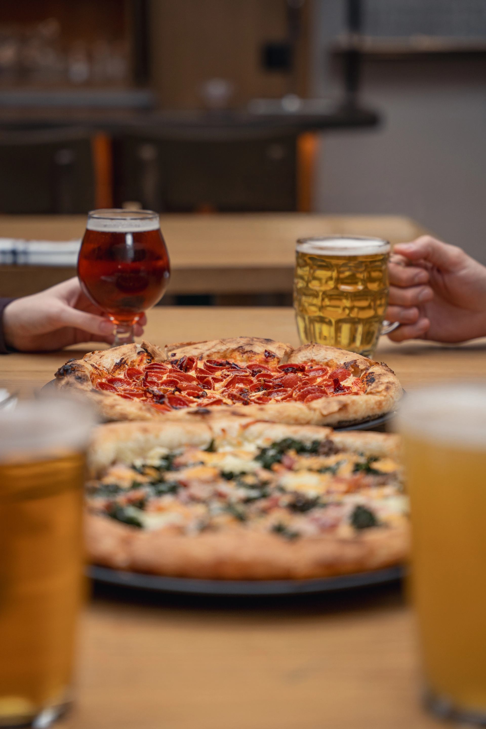 Hands holding glasses of beer next to two pizzas on a wooden table.