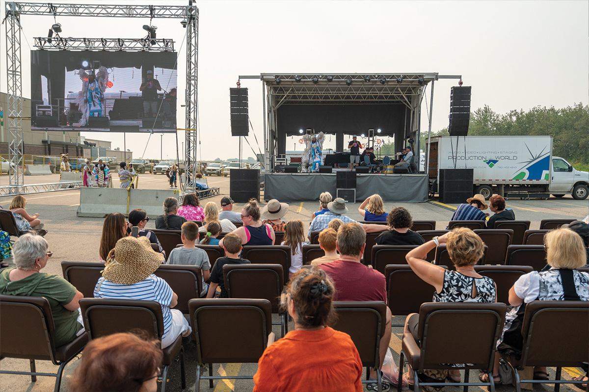 Audience sits outdoors watching performers on stage and large screen at community event.