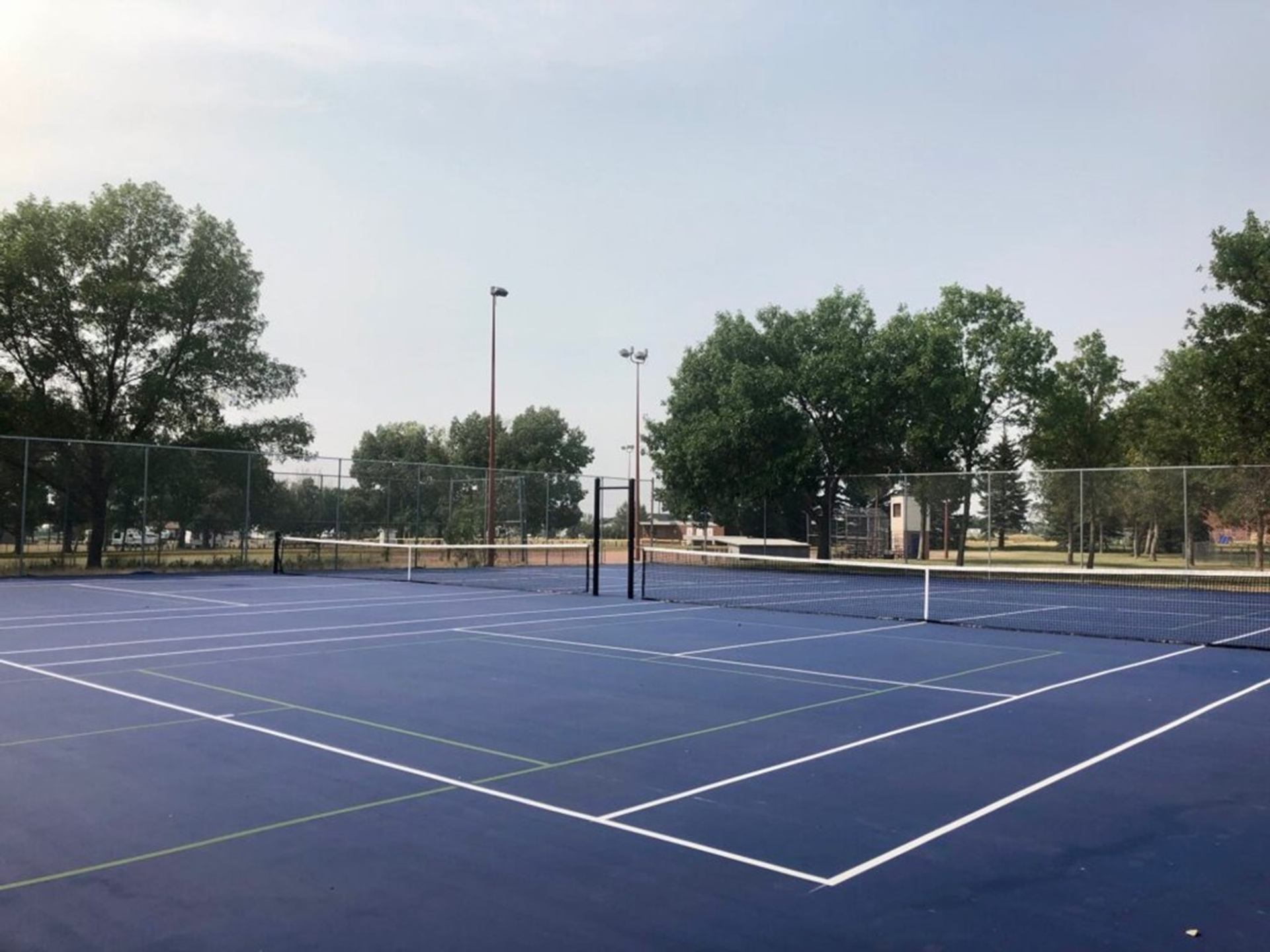 Empty blue tennis courts with white lines, surrounded by tall trees and chain-link fence.