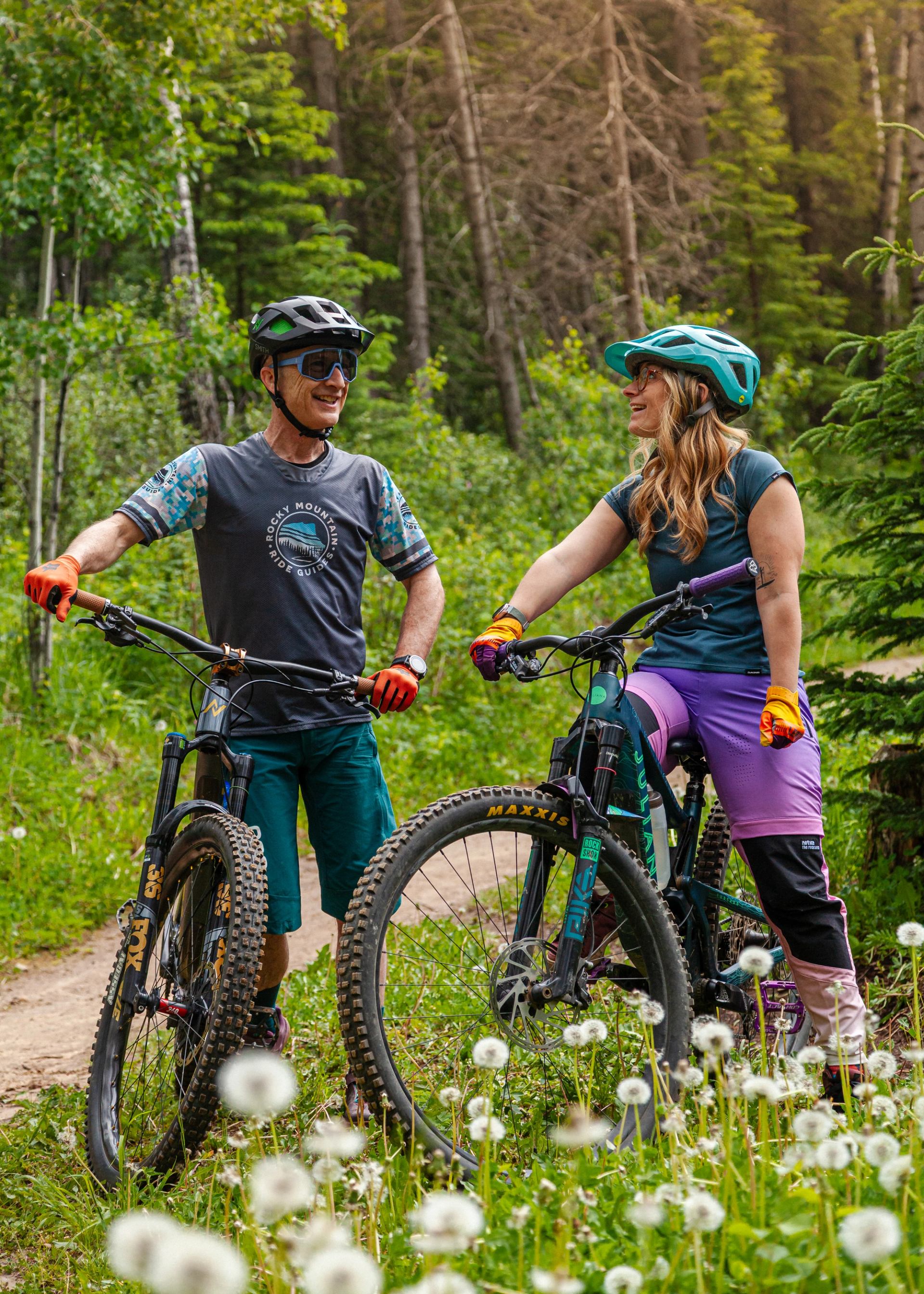 Two mountain bikers standing with bikes on a forest trail surrounded by wildflowers.