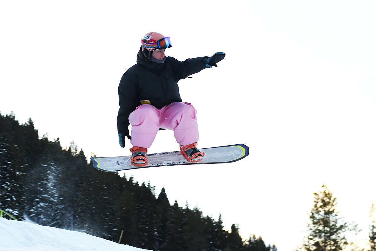 Ski snowboarder airborne on a snow-covered slope with trees in background.