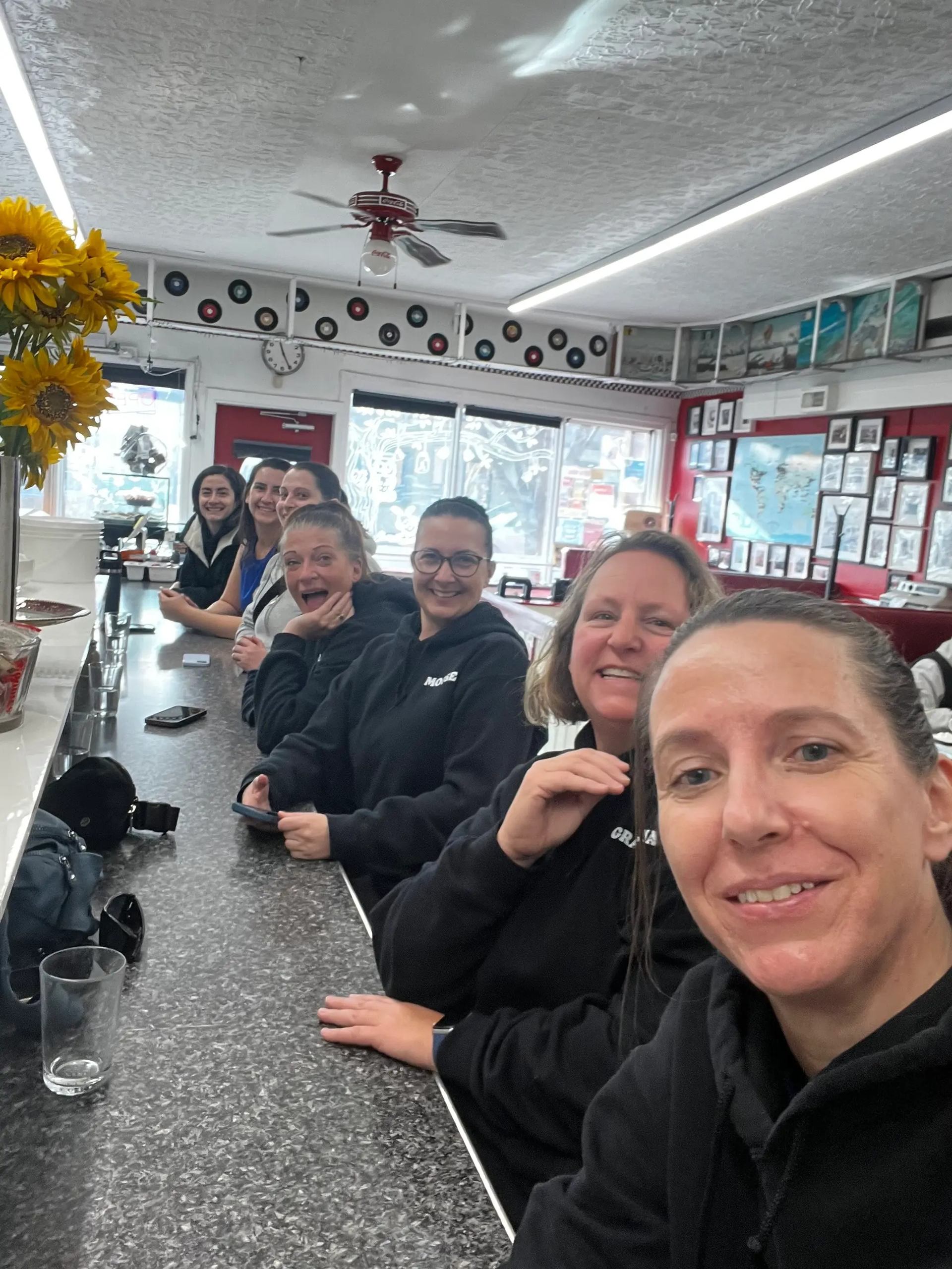 People sitting along a diner counter, smiling toward the camera amid retro decor.