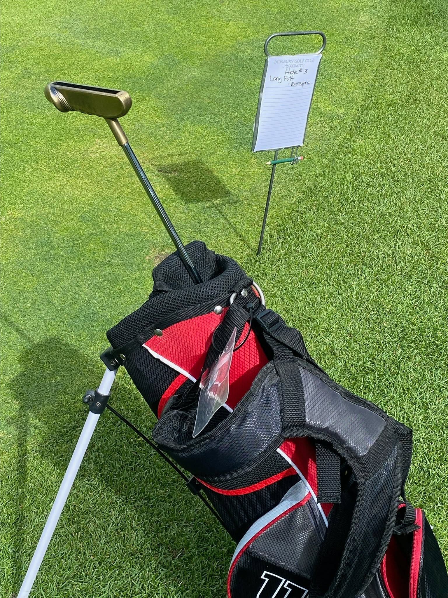 Golf bag with putter on green beside clipboard sign for long putt contest.