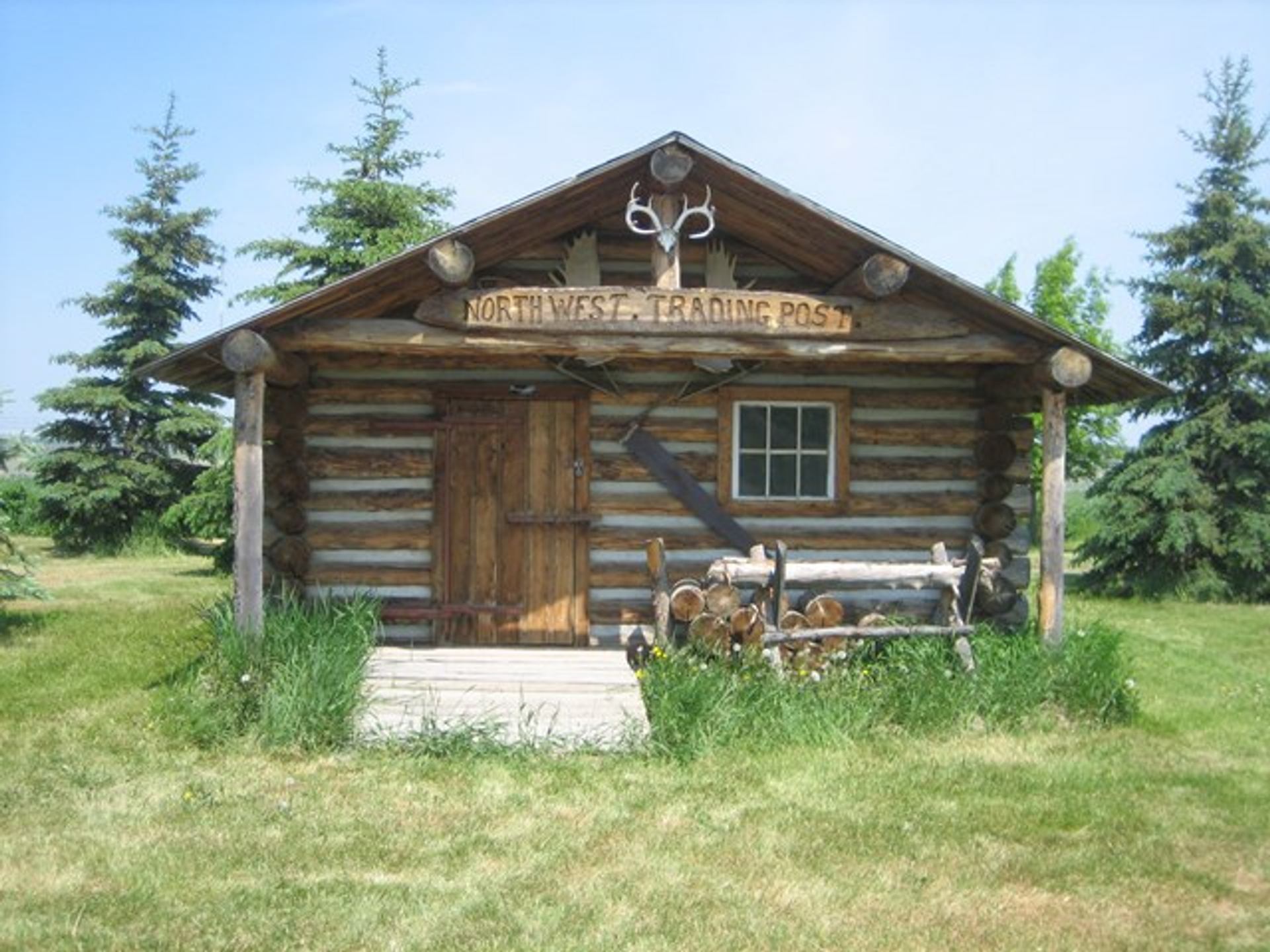 Log cabin labeled "North West Trading Post" with antlers above the sign. Surrounded by green grass and pine trees under a clear blue sky.