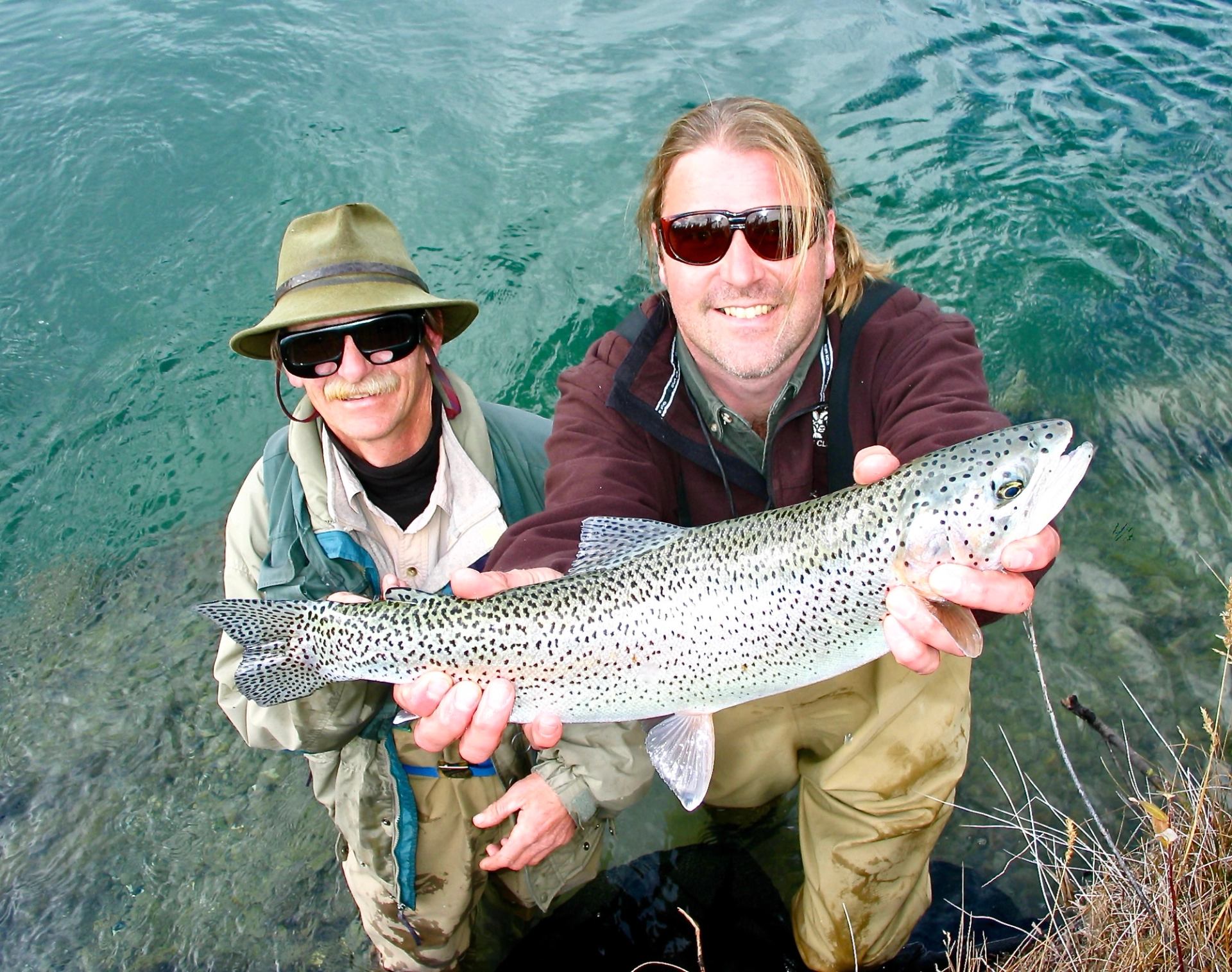 Two anglers in waders stand in clear water, holding a large speckled trout.