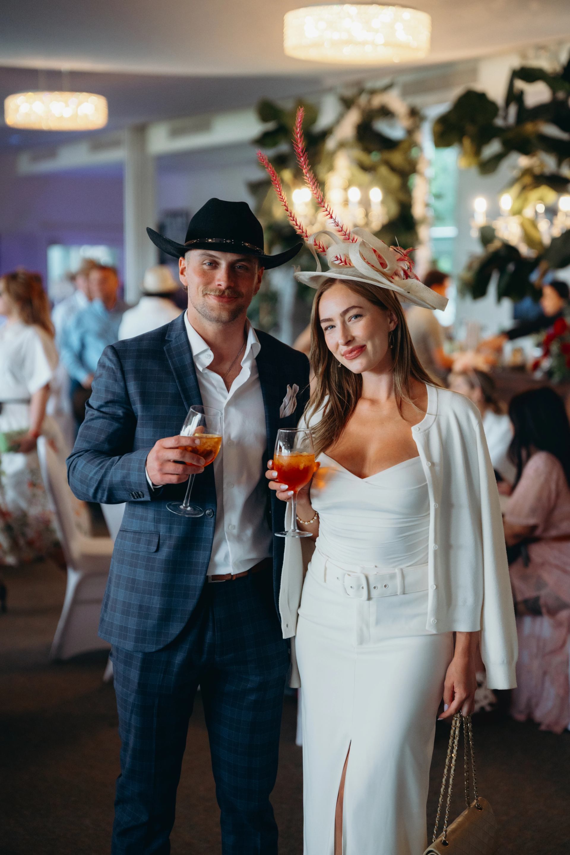 Two attendees dressed in formal race-day attire holding drinks at the event.