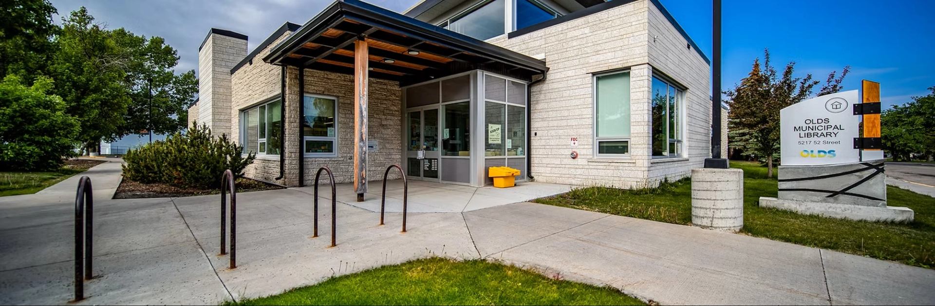 Olds Municipal Library exterior with glass entrance, bike racks, sidewalk and lawn.