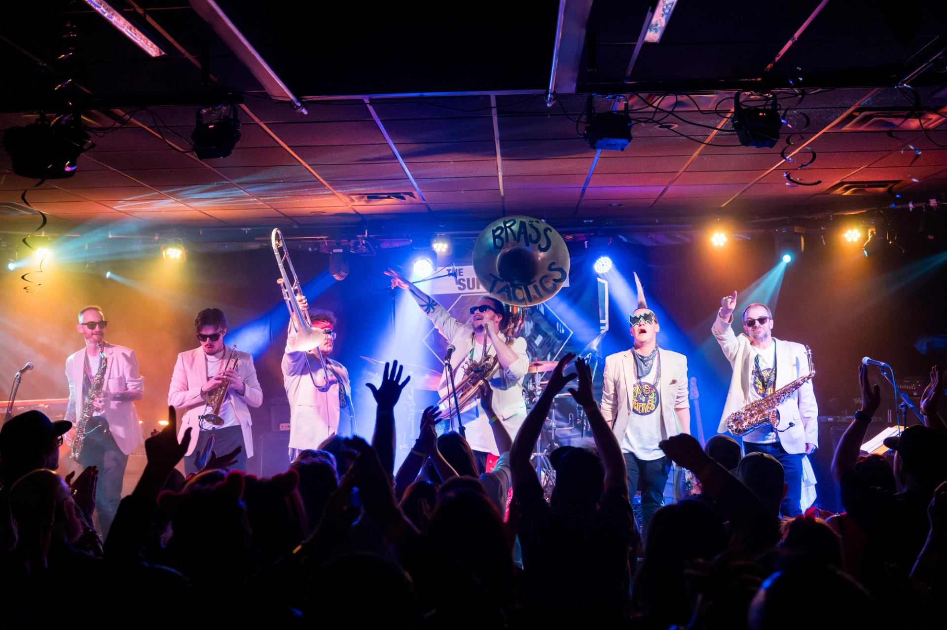 Brass band performs on colourful stage as crowd raises hands and silhouettes cheer.