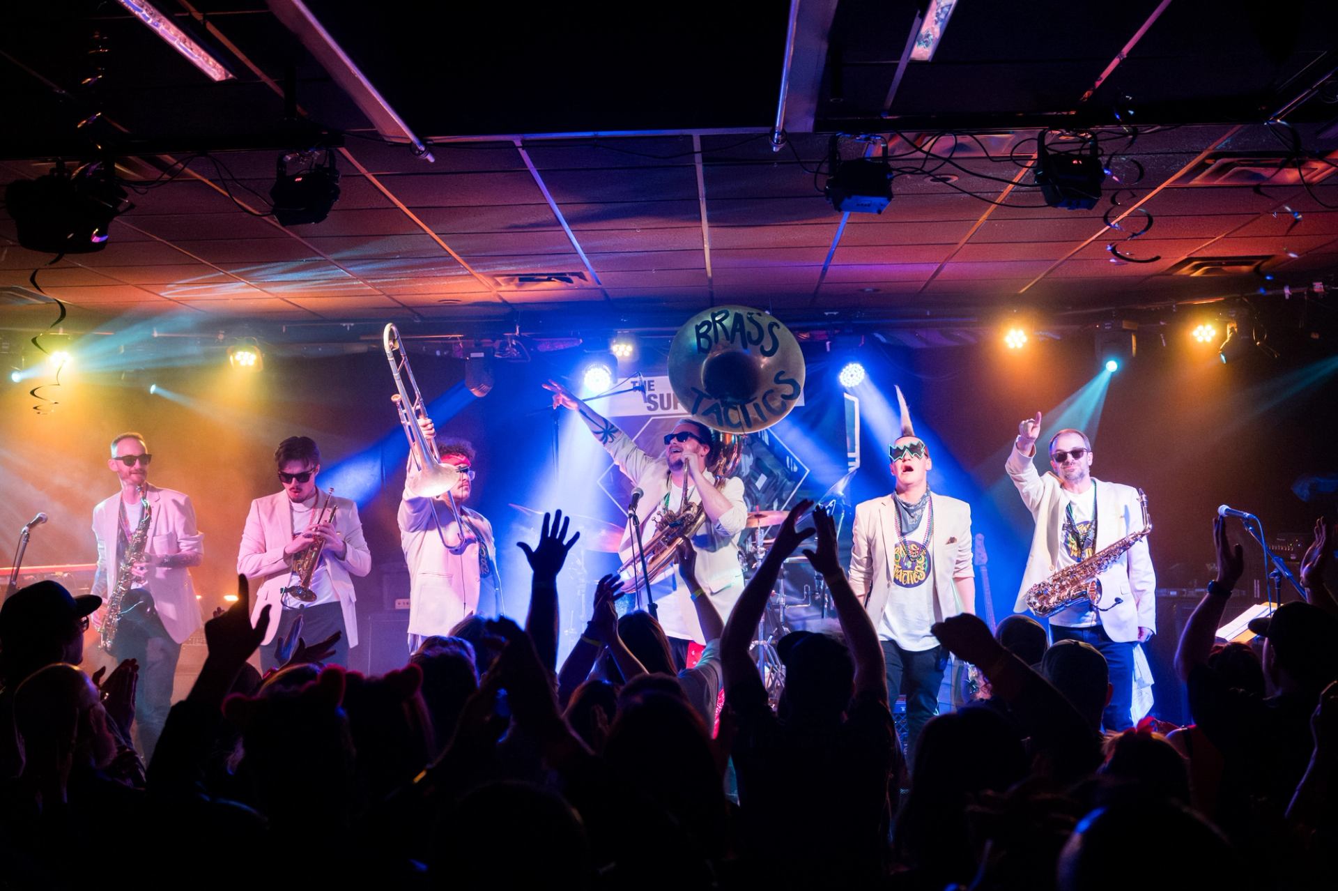 Brass band performs on colourful stage as crowd raises hands and silhouettes cheer.