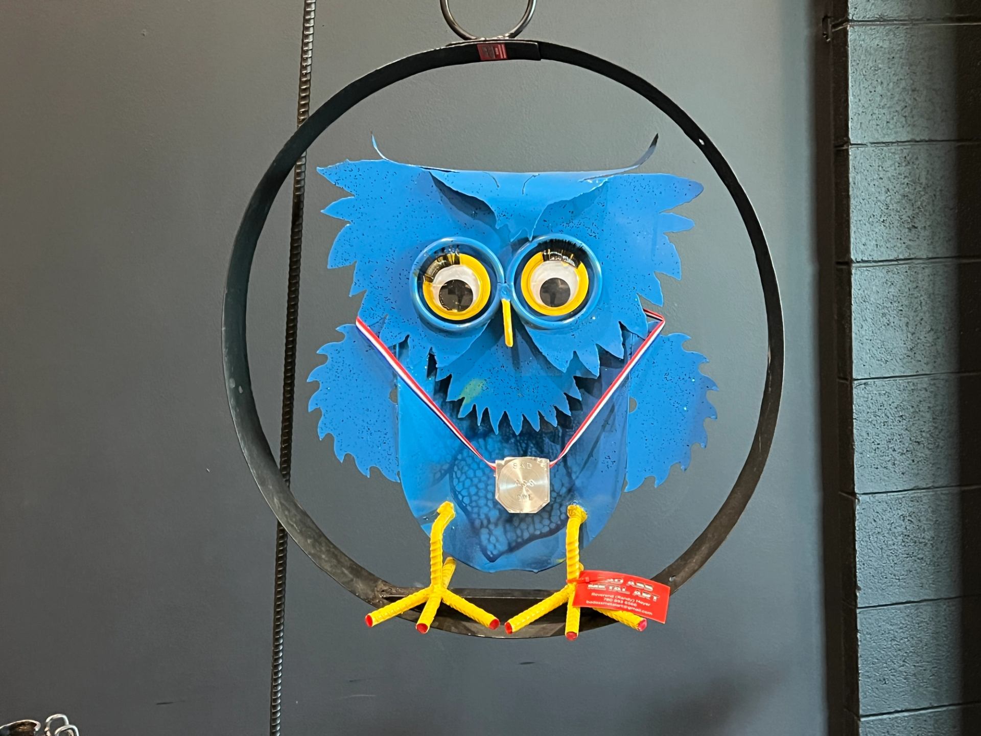 Blue metal owl sculpture hangs in black ring, medal ribbon and red tag.
