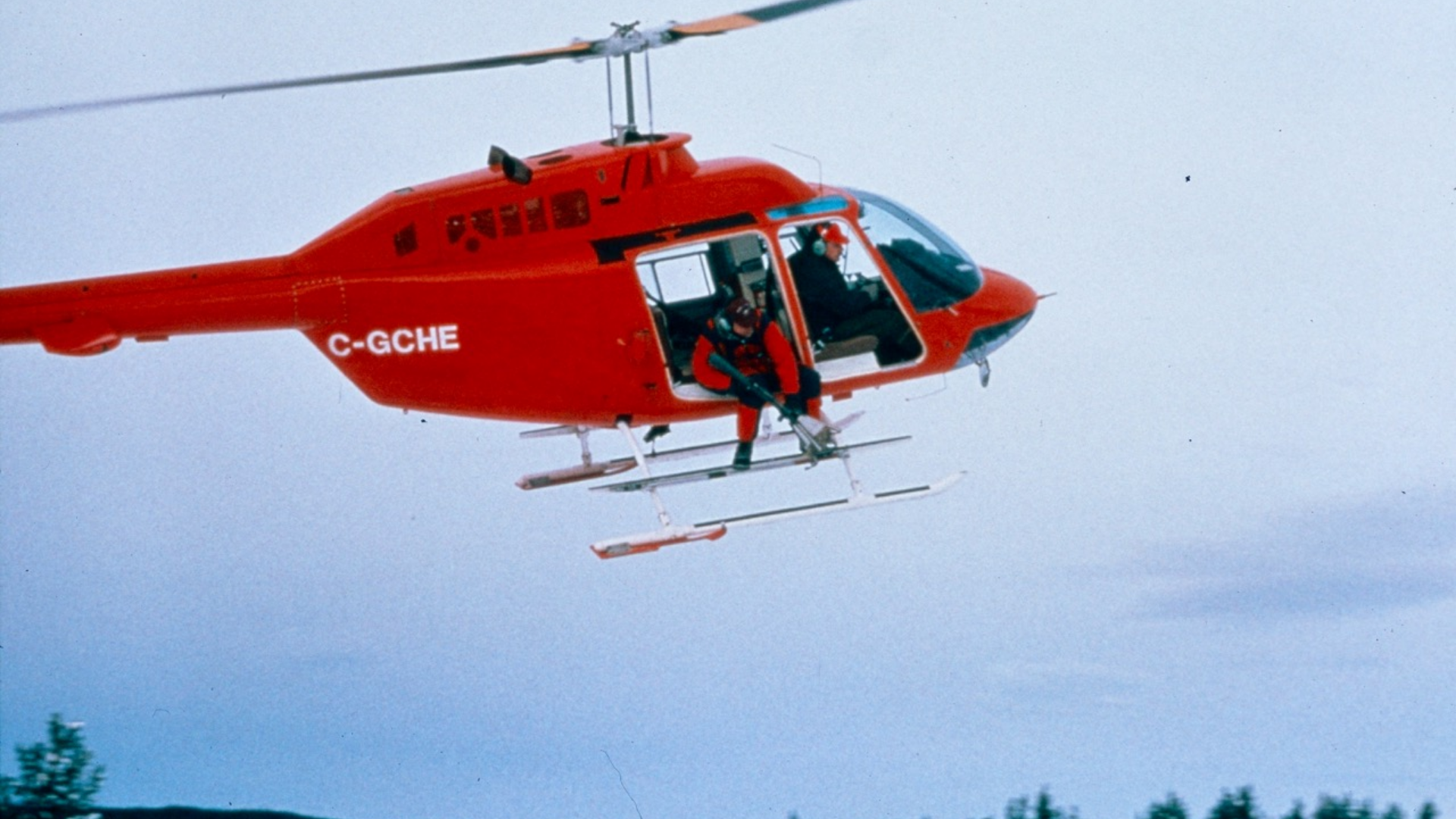 Red helicopter in flight with open door and person sitting on landing skid.