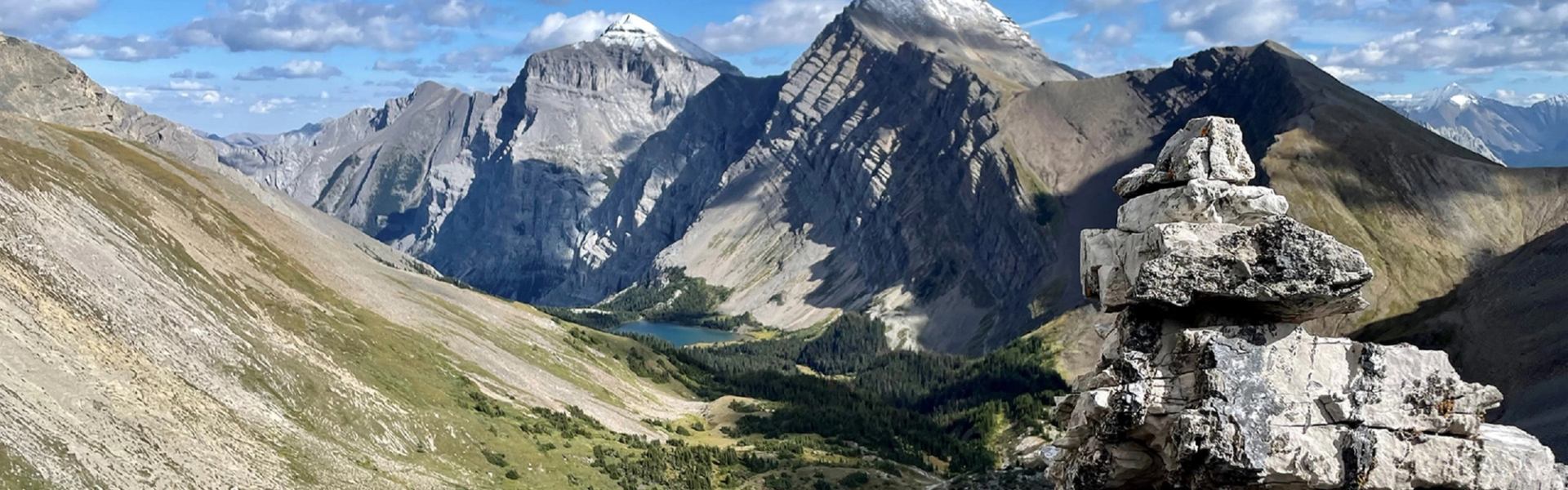 Rock cairn overlooks steep mountain valley with alpine lake and rugged peaks.