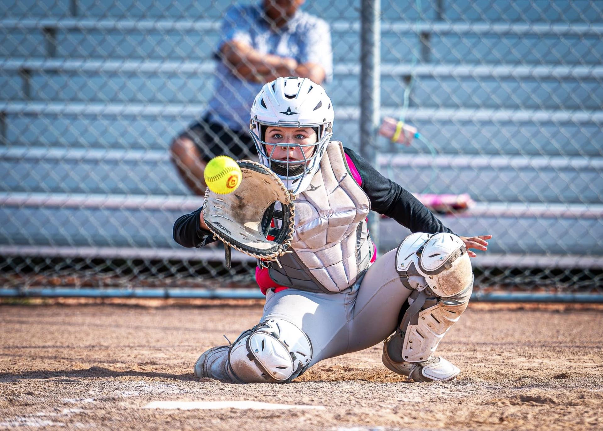 Softball catcher in full gear kneels in dirt, glove reaching toward incoming ball.