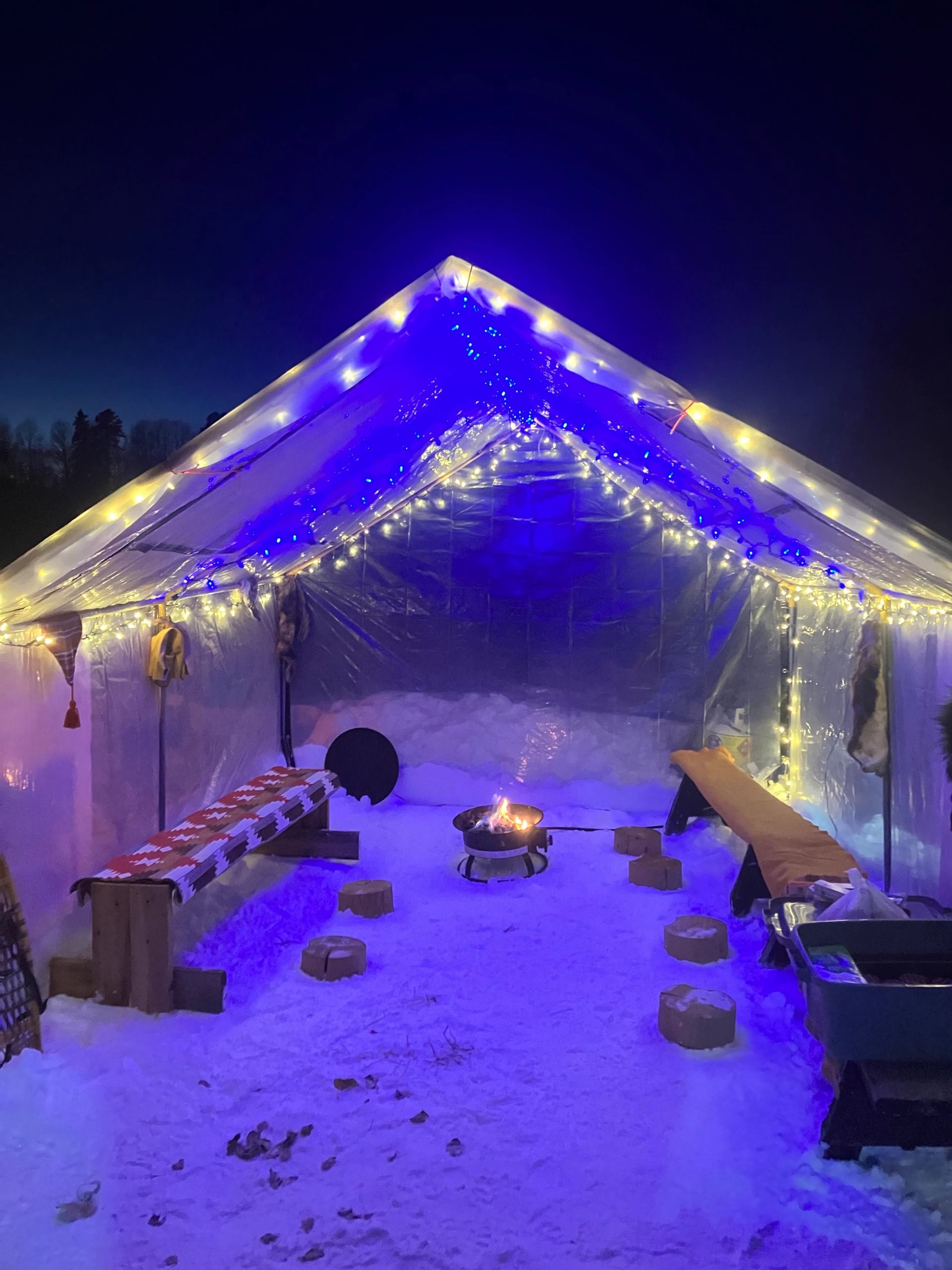 Clear tent lit with string lights and a fire pit inside surrounded by snow