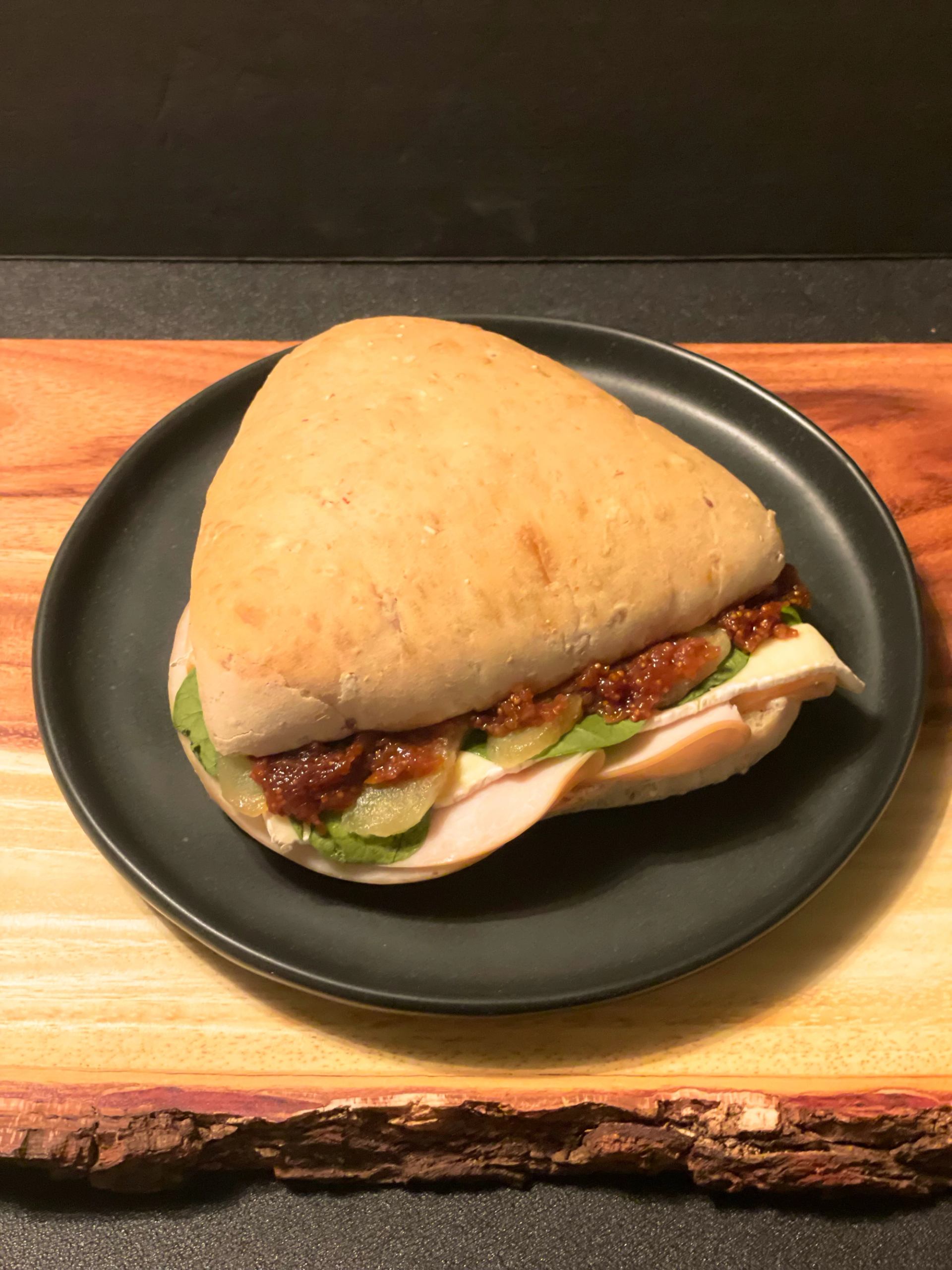Triangle sandwich with meat, greens and relish on black plate over wooden board.