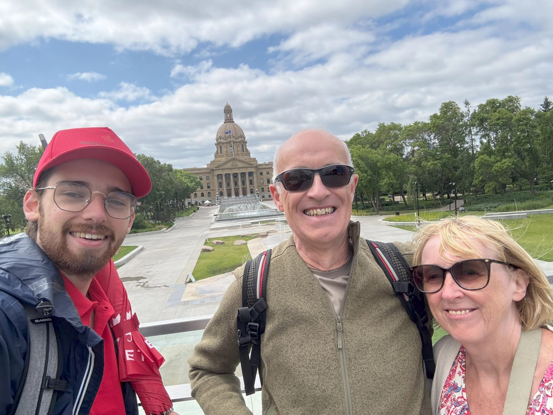Three people stand near a domed landmark with fountain and greenery in Edmonton.