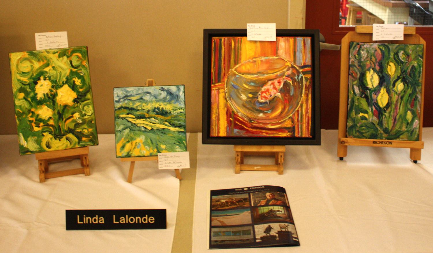 Art display table with four colourful paintings on easels and a Linda Lalonde nameplate.
