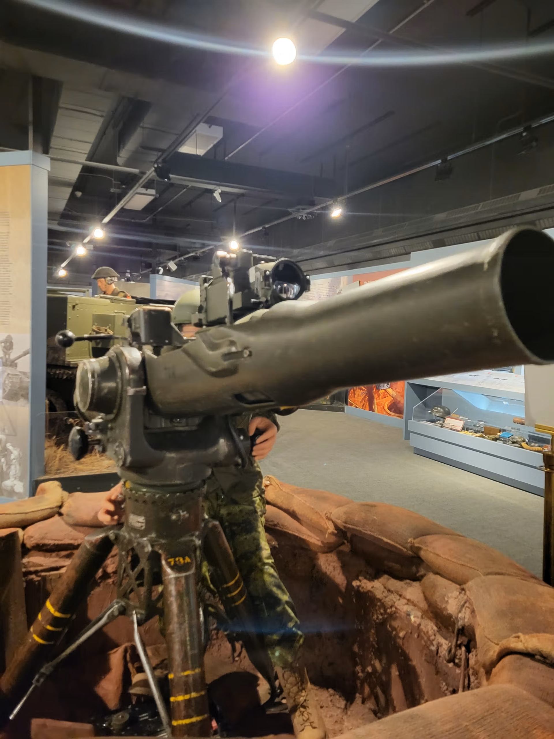 A large military weapon on a tripod, set in a sandbagged trench exhibit in a museum.