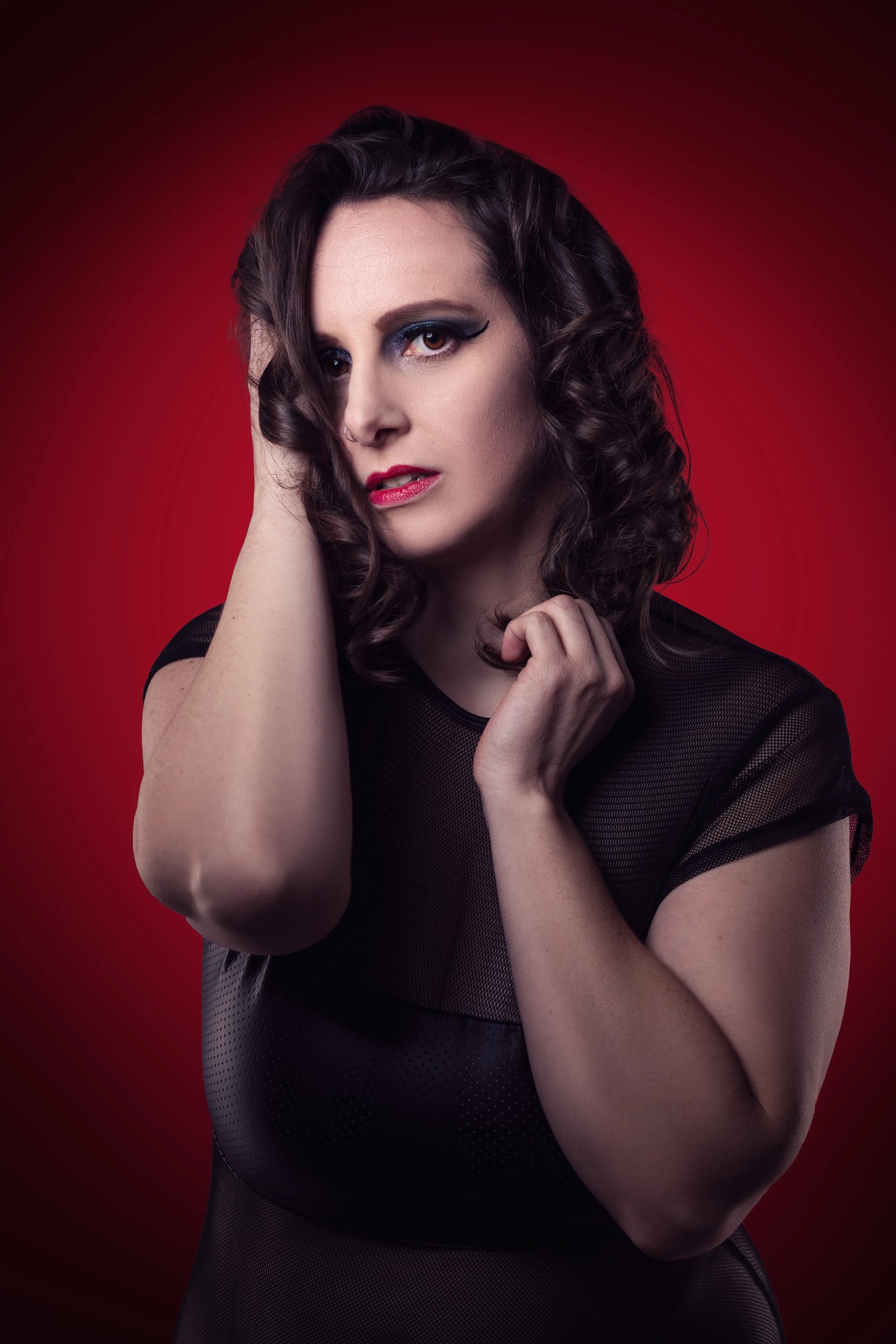 A woman with dark curly hair, dramatic blue winged eyeliner, and red lipstick, wearing a black mesh top, against a red background.