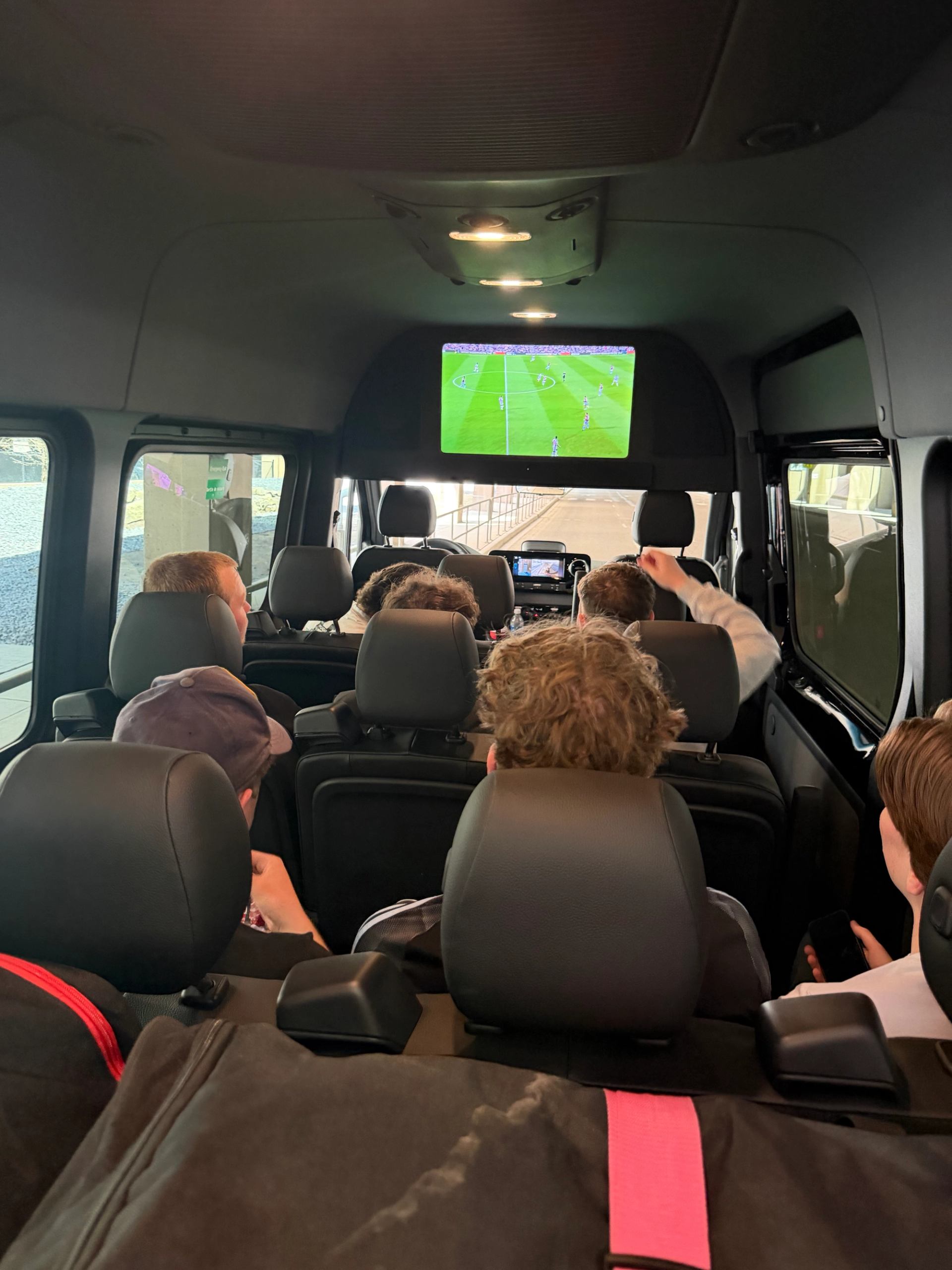 Passengers sit in a van watching a soccer match on an overhead screen.
