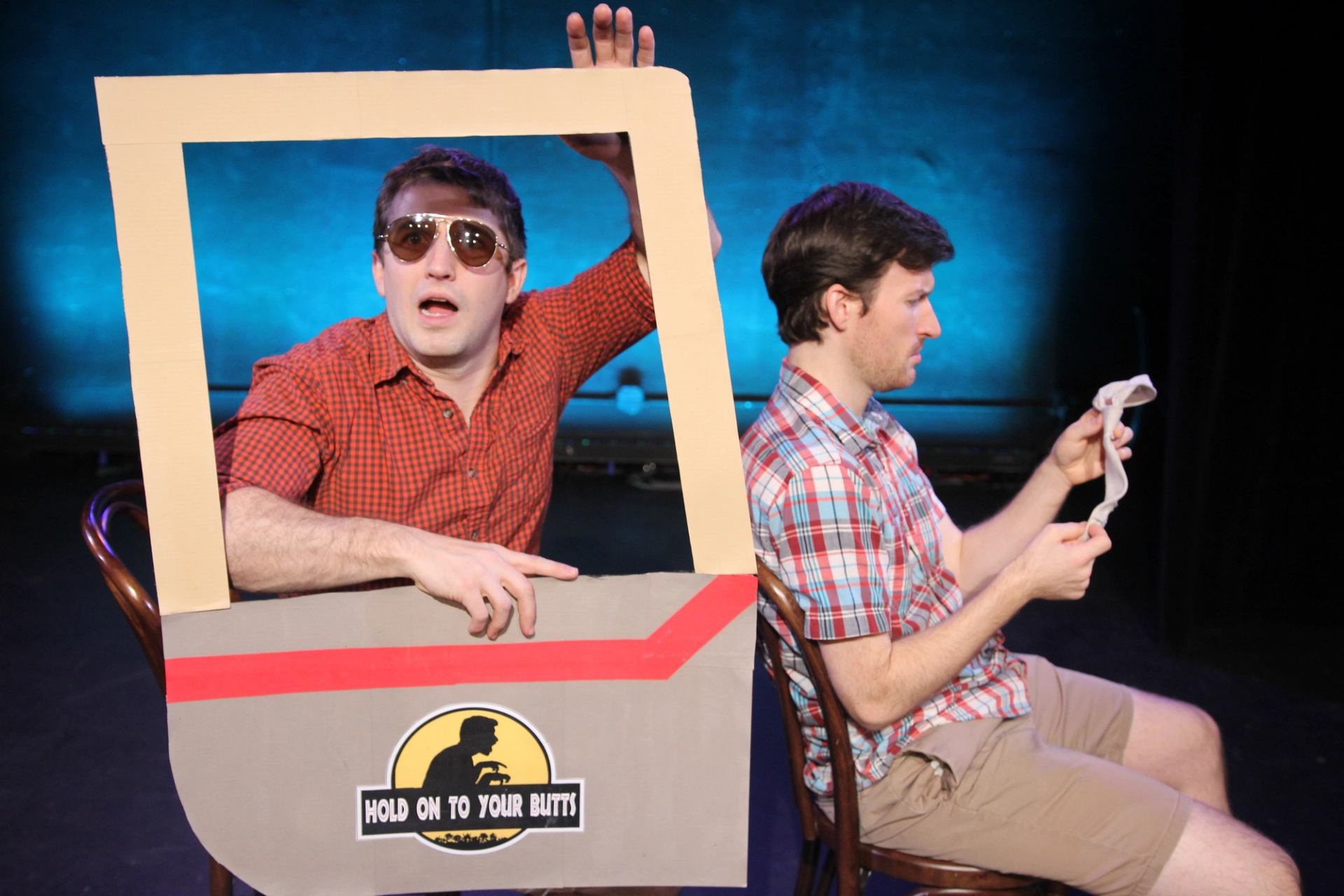 Stage performers sit in cardboard car prop with “Hold On To Your Butts” sign.