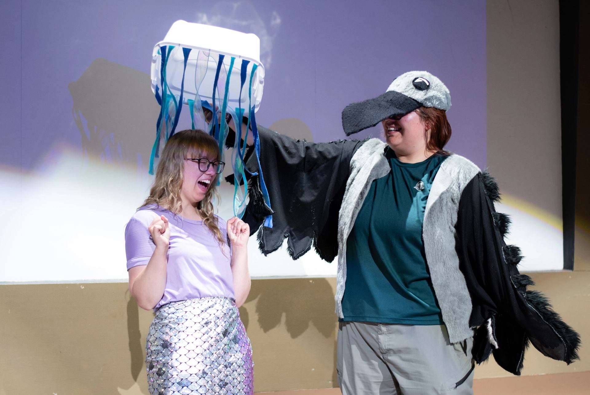Theatre performers in costumes hold a blue and white draped prop on stage.