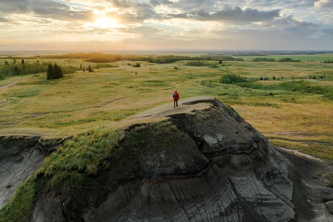 Person walking on a rocky hilltop above a wide prairie landscape under a cloudy sky.