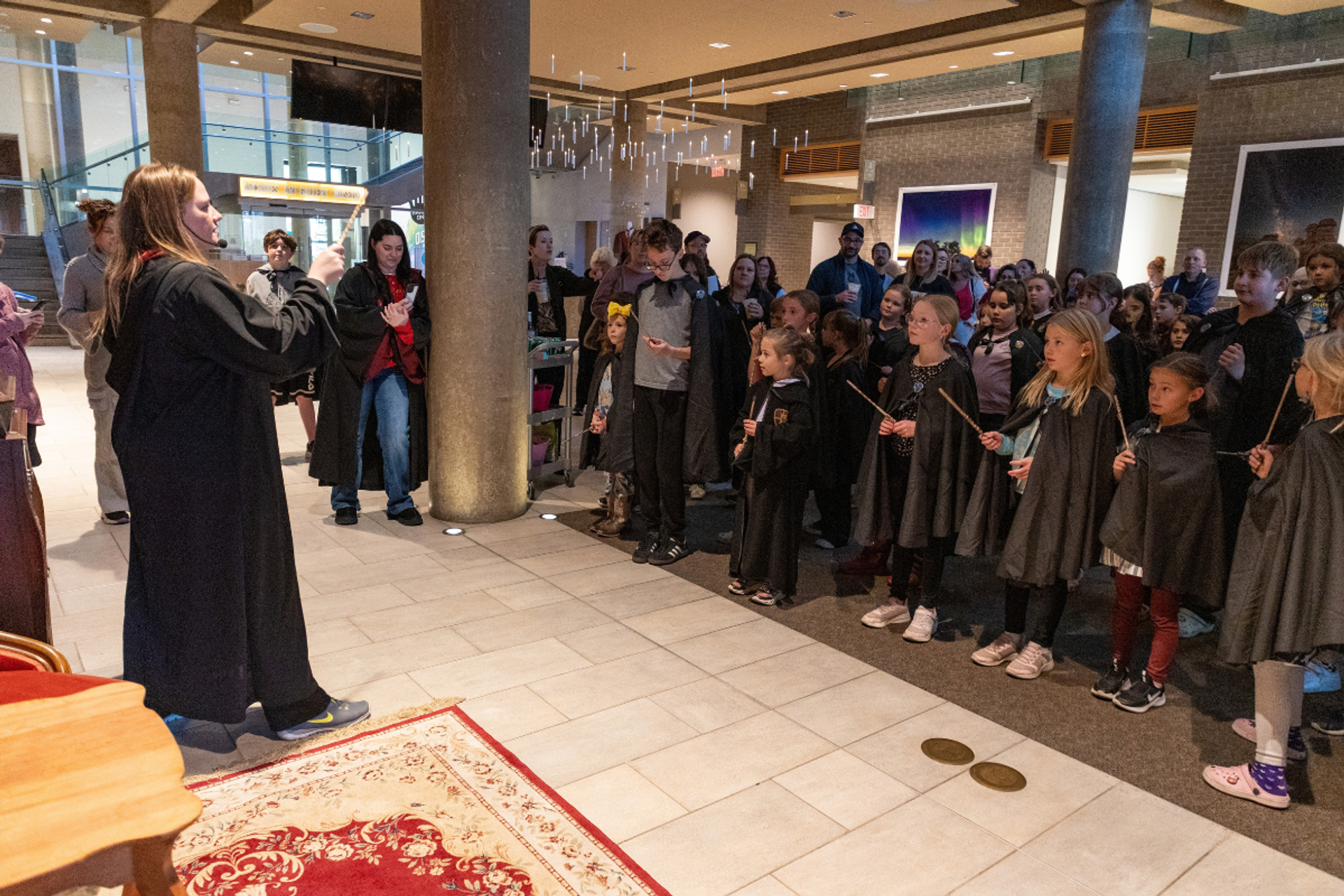 Costumed performer addresses children holding wands in a crowded indoor lobby.