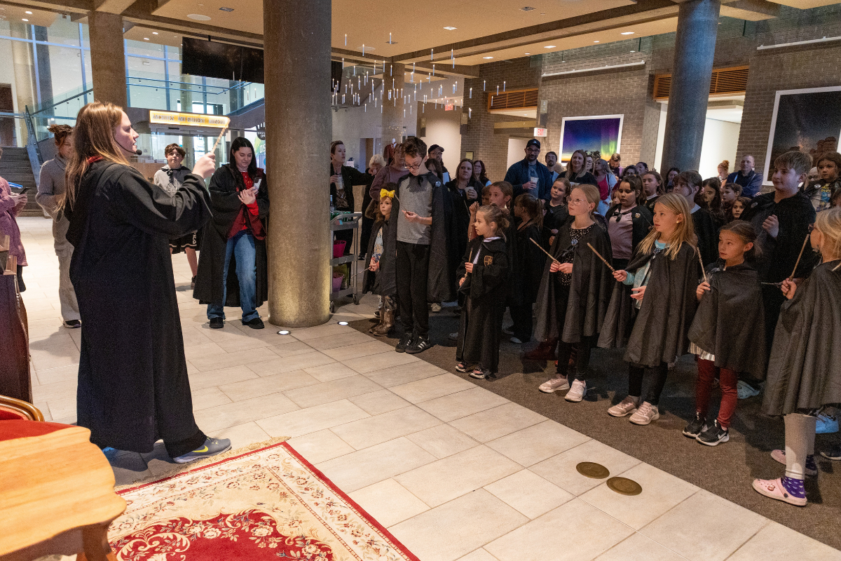 Costumed performer addresses children holding wands in a crowded indoor lobby.