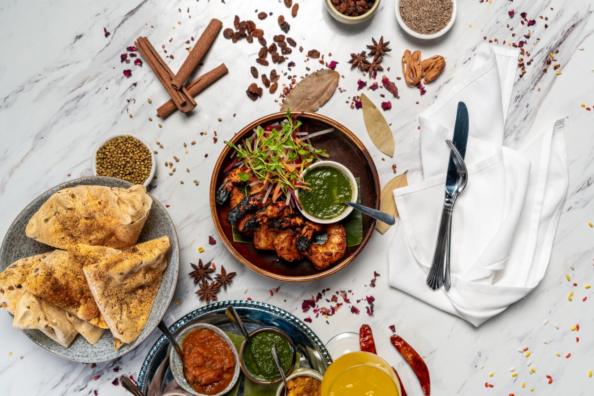 Grilled shrimp with green chutney, flatbread, chutneys and spices on marble tabletop.