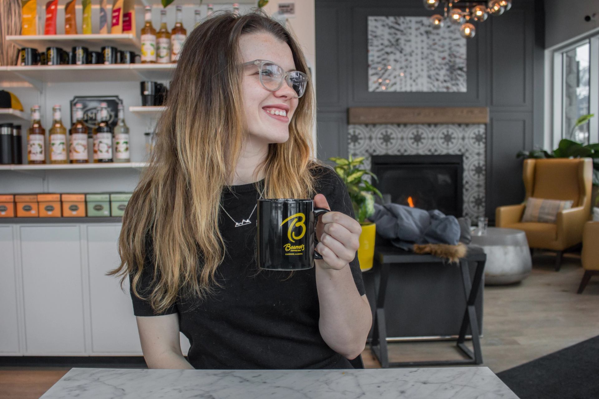 Person holding a Beamer’s Coffee Bar mug while sitting at a table near a fireplace in a café.