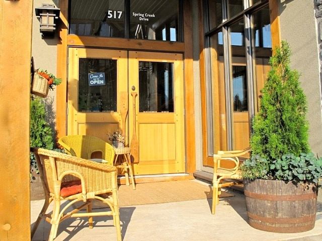 Gallery entrance with wooden double doors, wicker chairs, and potted greenery in sunlight.