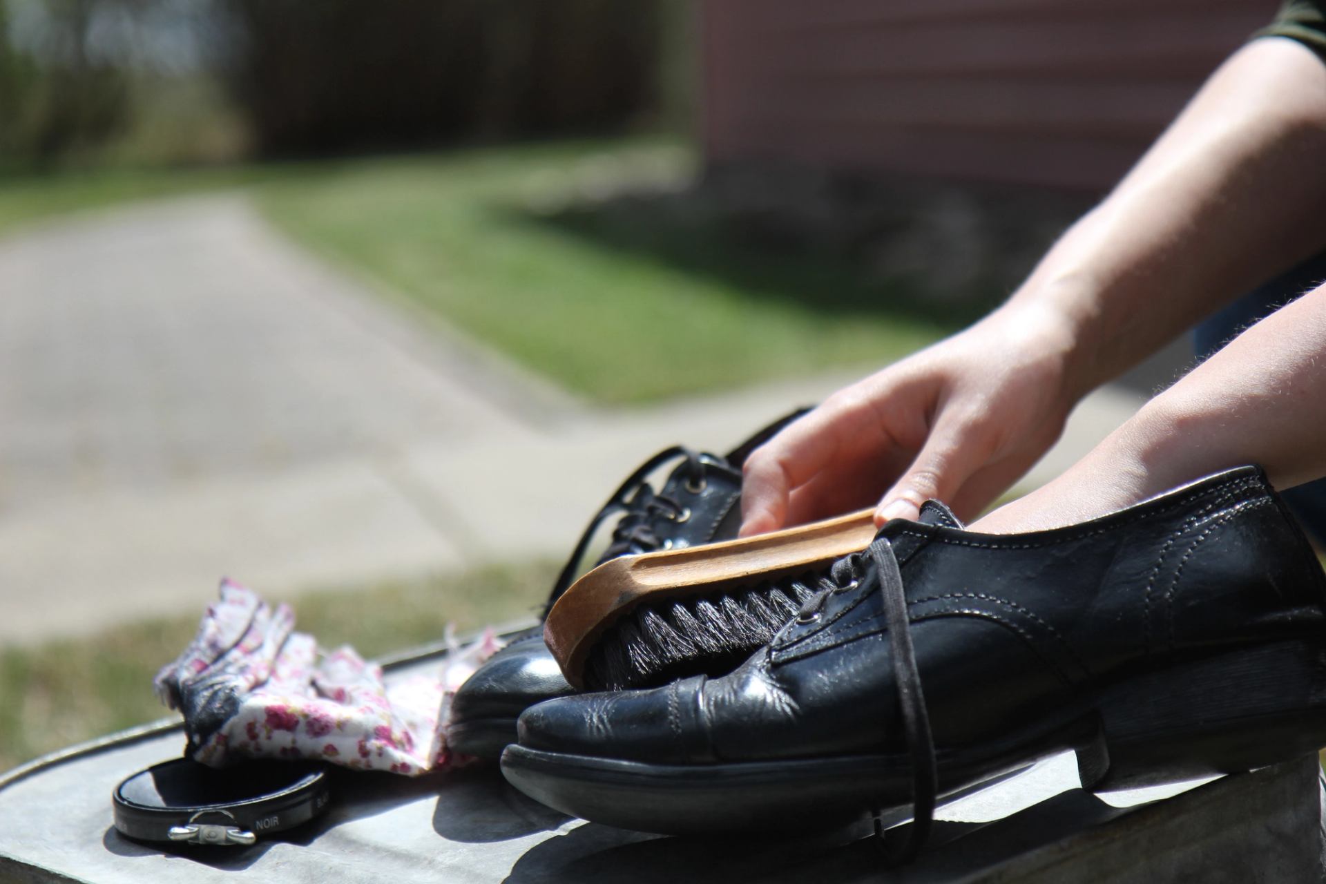 Hands polishing black leather shoes with a brush on an outdoor surface.