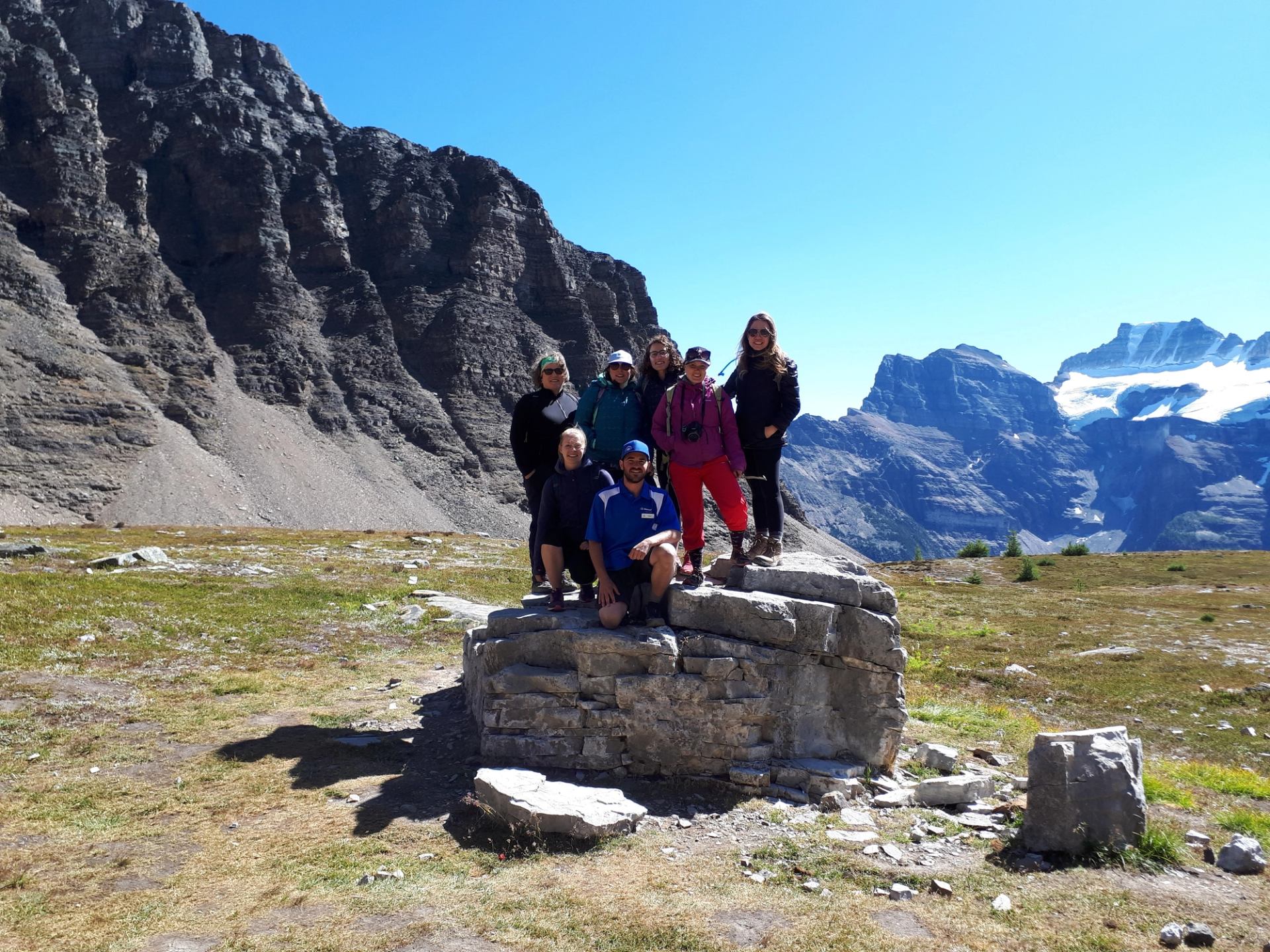 Hikers stand on a stone ruin in a rocky alpine meadow with mountains.