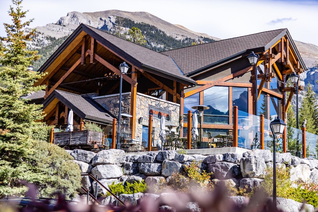 Rustic stone and timber building set against tall evergreen trees and rocky mountain peaks.