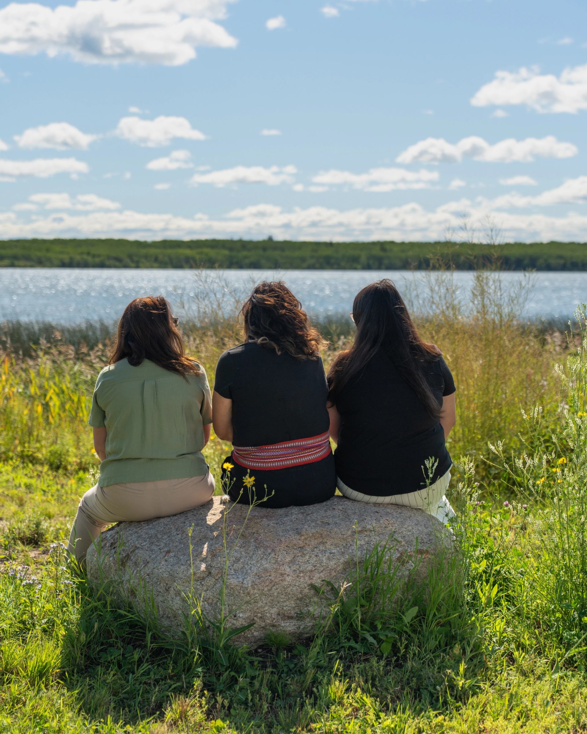 People sit on a boulder beside a lake, looking across water.