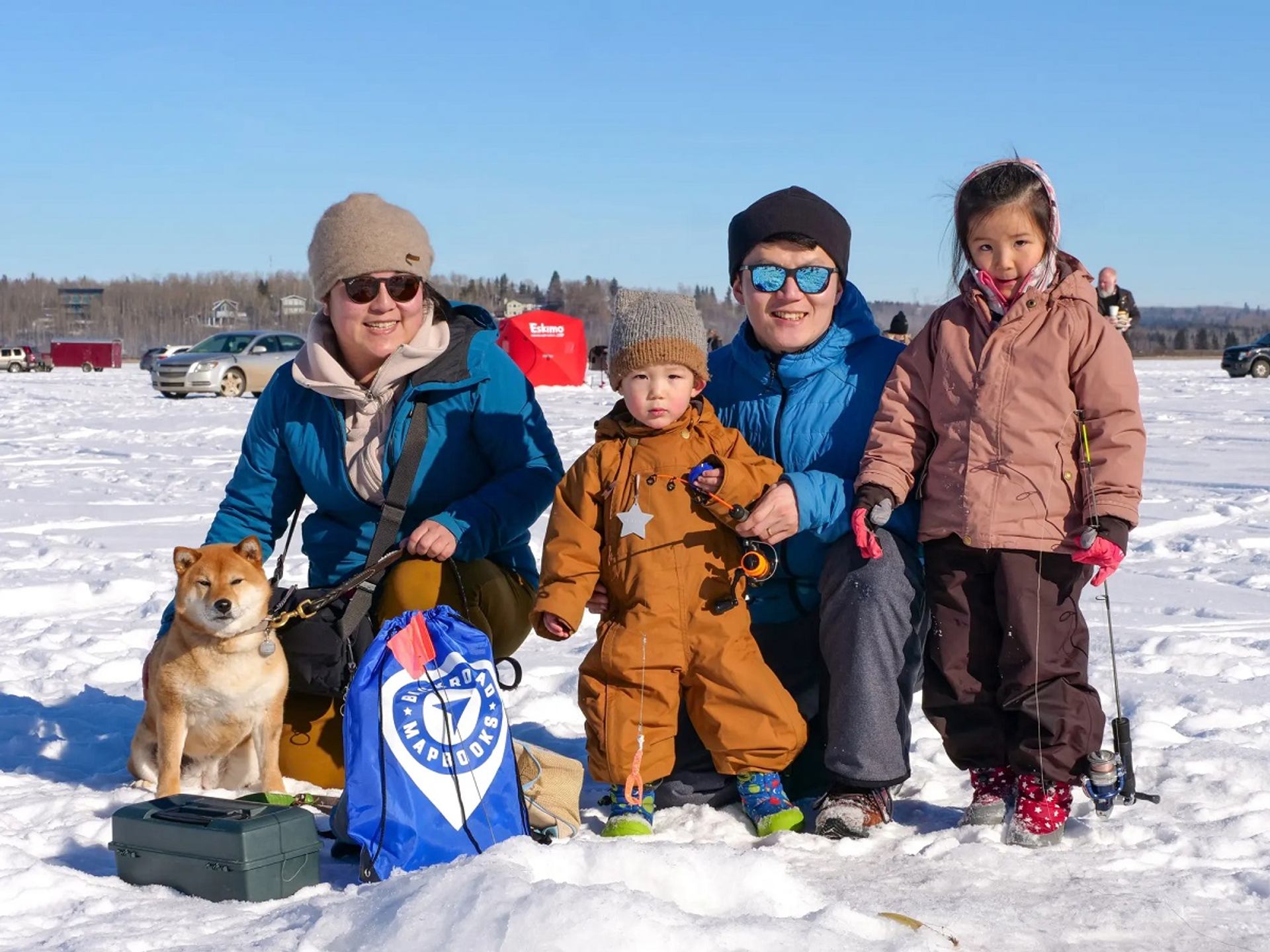 Group of people and a dog on snowy ice with fishing gear and vehicles.