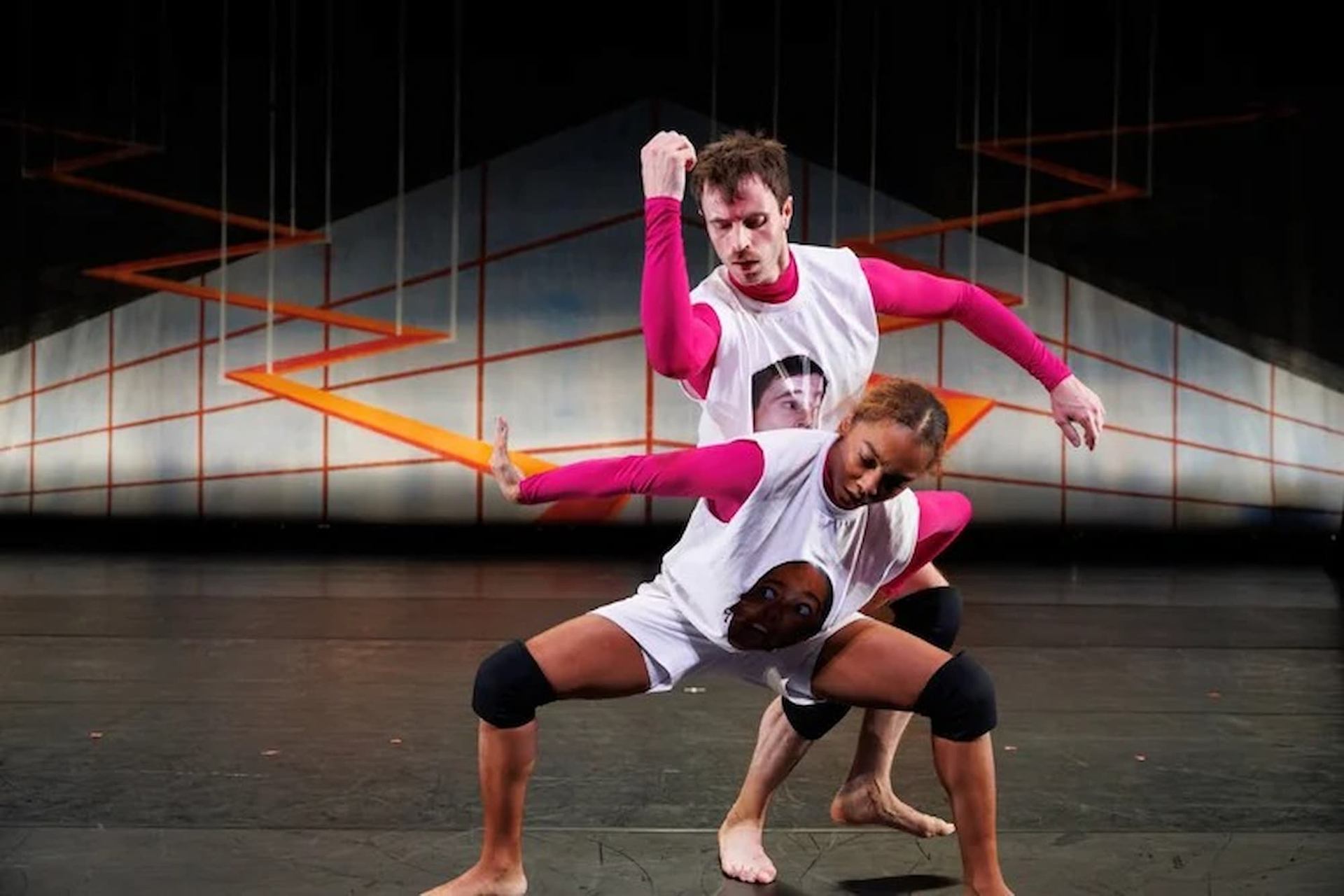 Two contemporary dancers in white shirts and pink sleeves pose onstage before geometric backdrop.