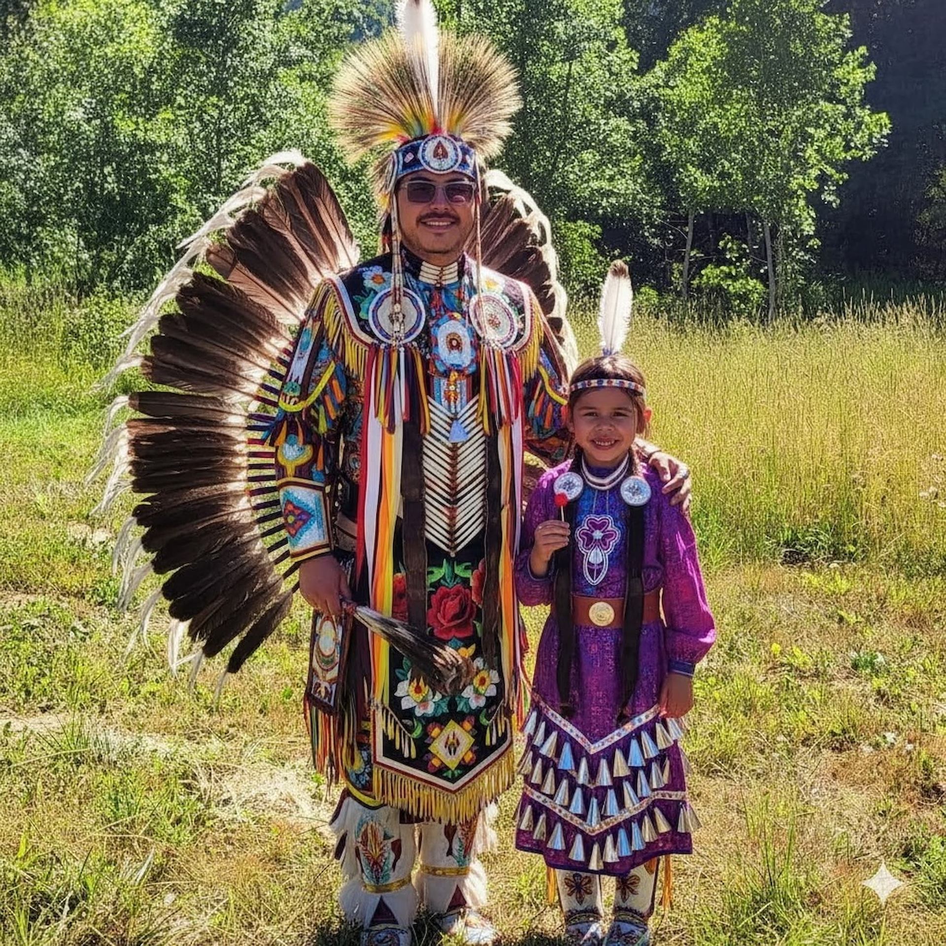 Two people in colourful traditional regalia stand in a grassy field with trees.