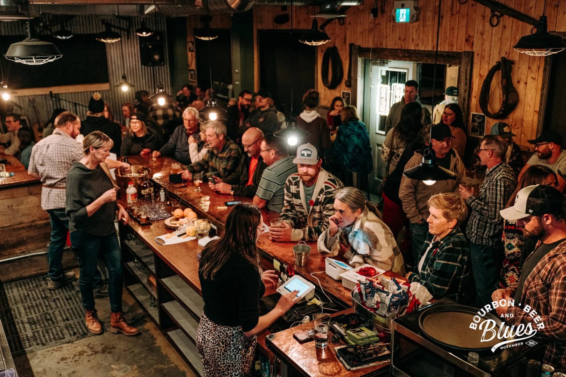 A lively crowd of people gathered around a long wooden bar, with bartenders serving drinks in a rustic setting.