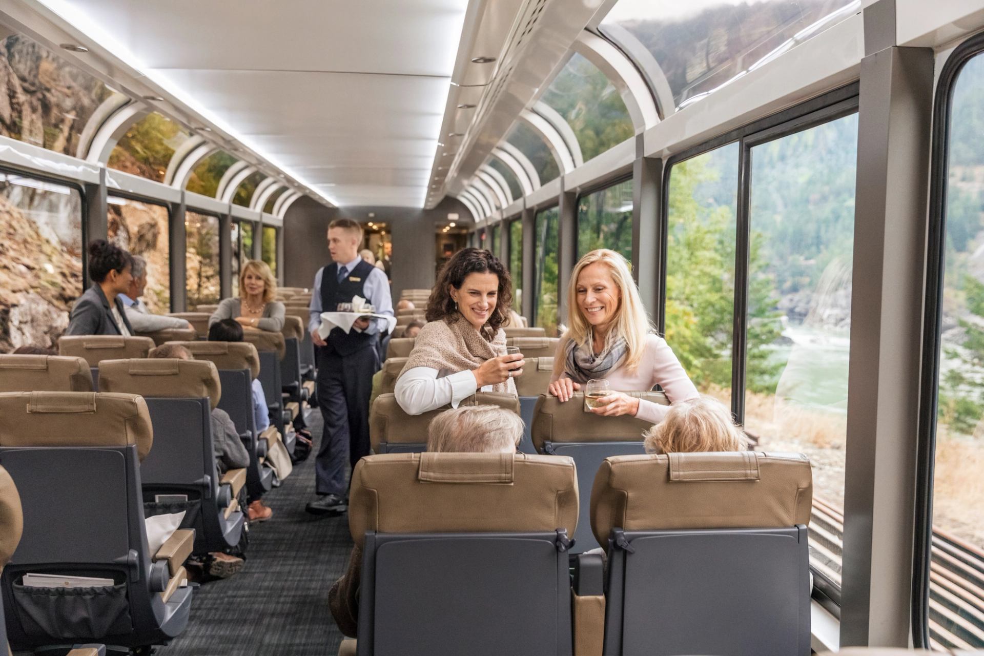 Rocky Mountaineer glass‑dome coach interior with wide seats, service aisle, and scenic river views outside.
