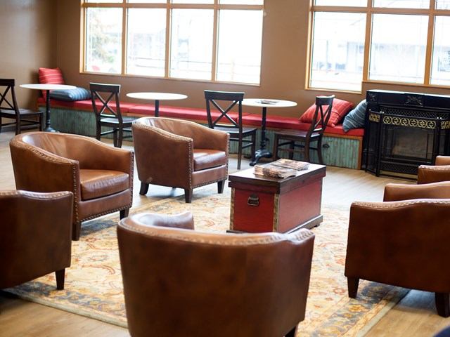 Cozy interior with armchairs around a vintage trunk table, cushioned benches along the walls, and large windows. Warm, inviting atmosphere.