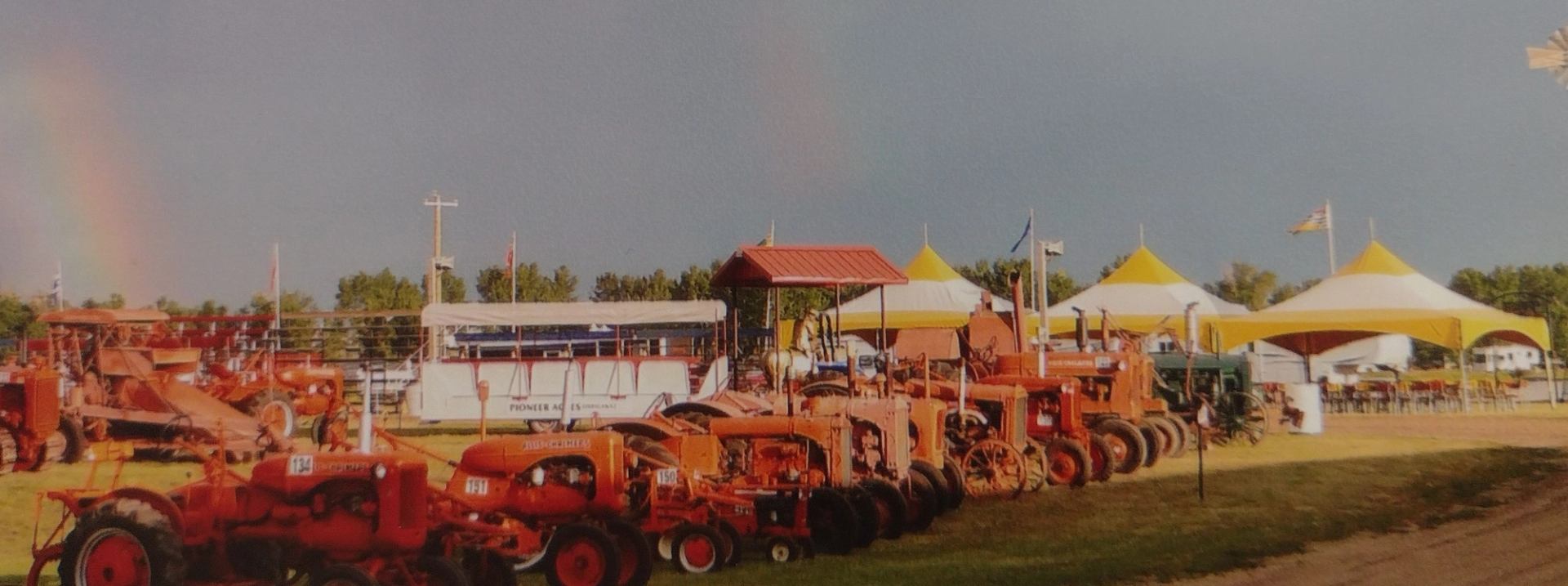 Farm machinery is lined up in front of white and yellow event tents.