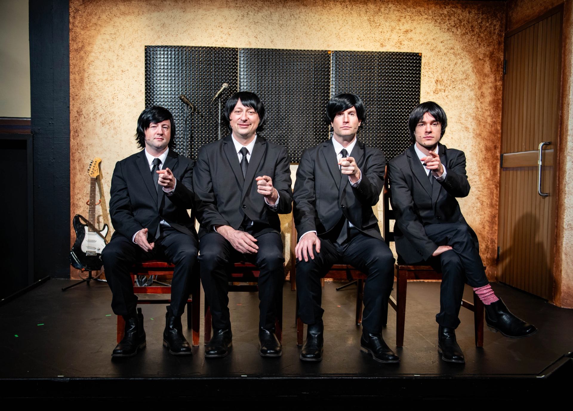 Four people in matching black suits sit on stage, pointing toward the camera.