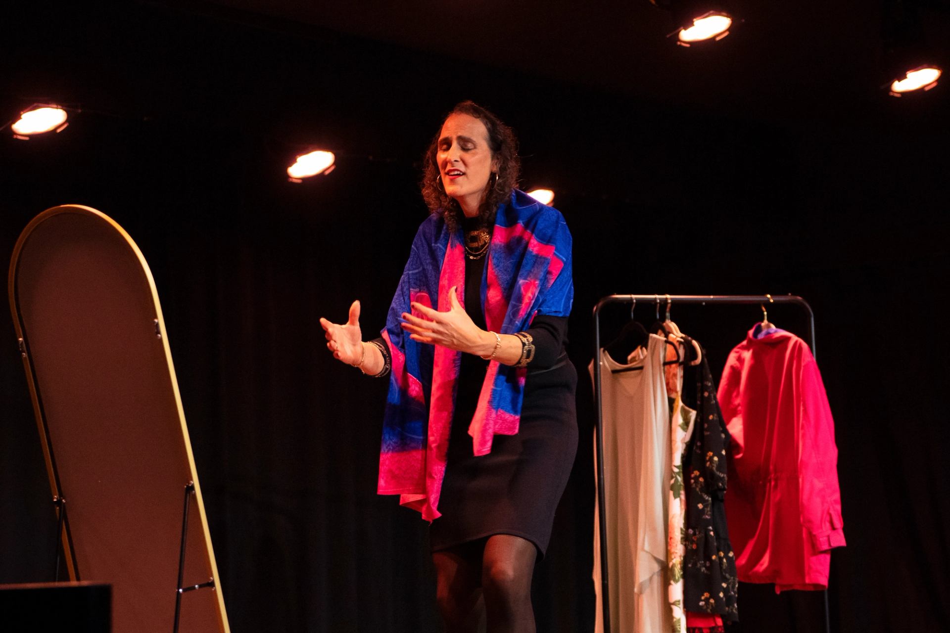 Performer on stage gestures with hands near clothing rack and standing mirror.