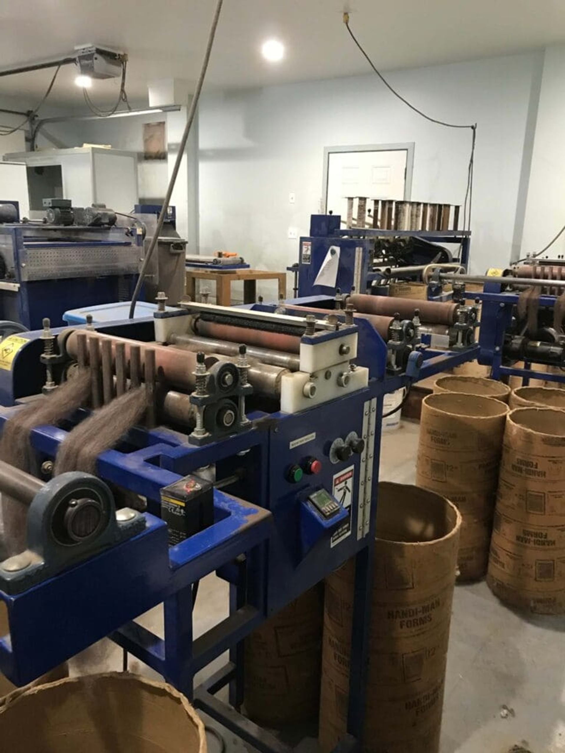 Industrial workshop with blue machinery feeding material into tall cardboard tubes on floor.