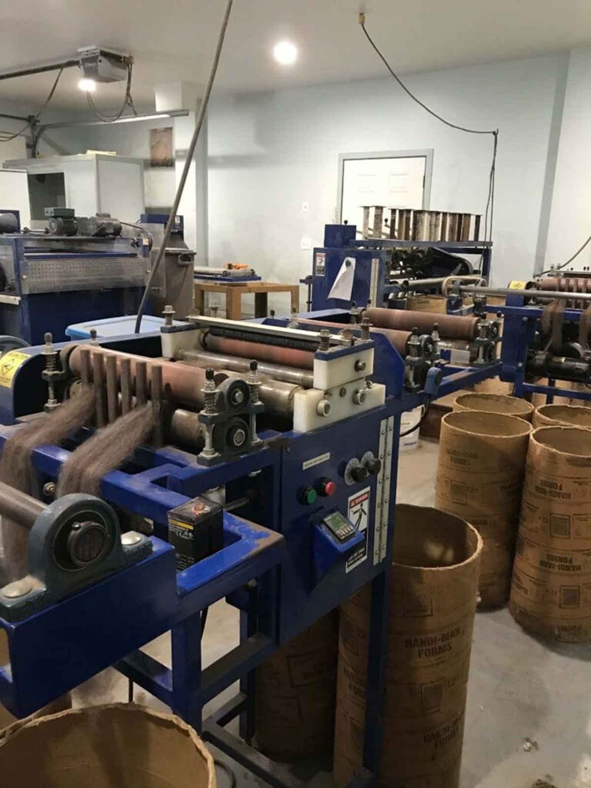 Industrial workshop with blue machinery feeding material into tall cardboard tubes on floor.