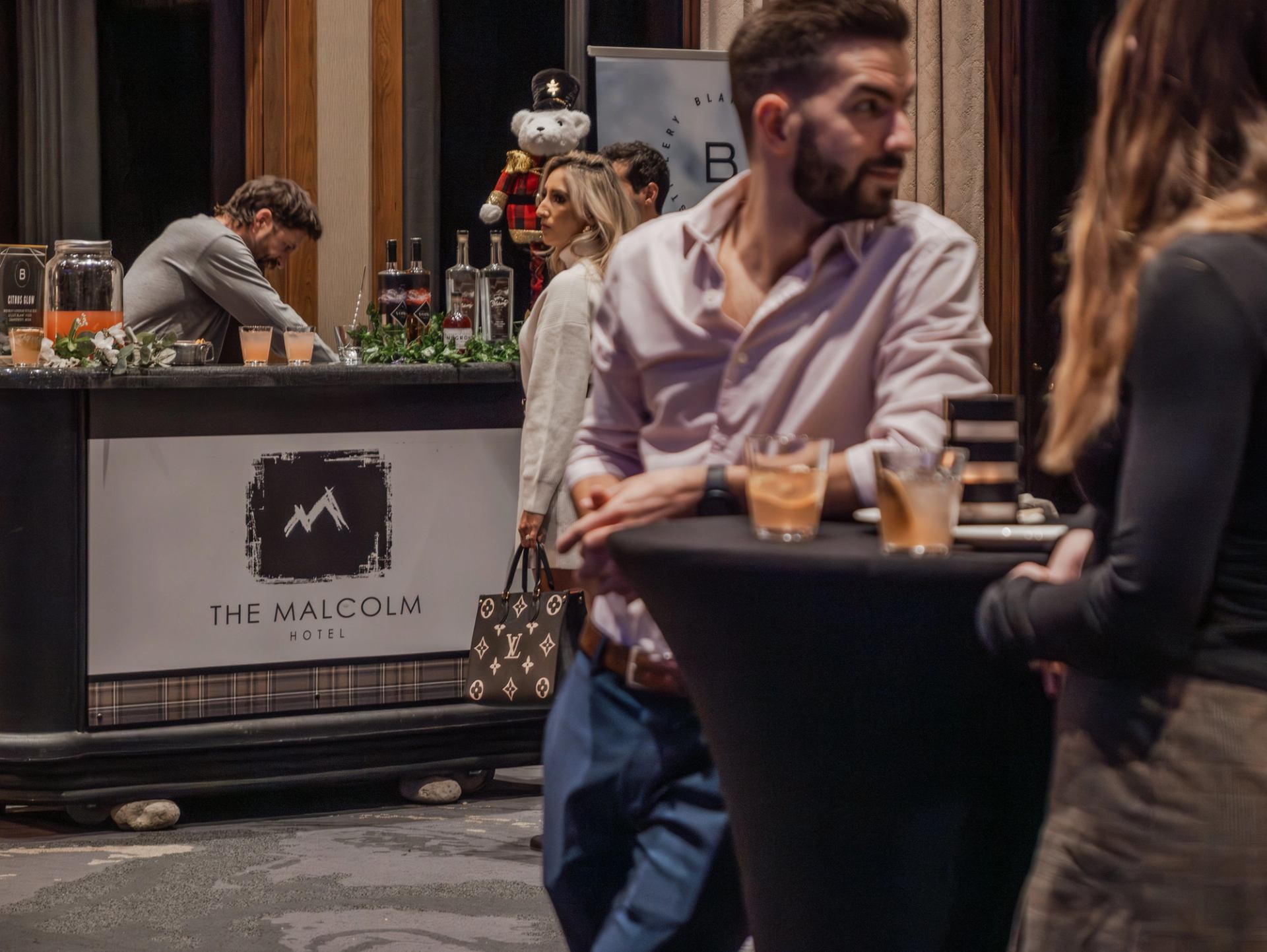 People standing around a cocktail table with drinks near a bar counter labeled “The Malcolm Hotel”